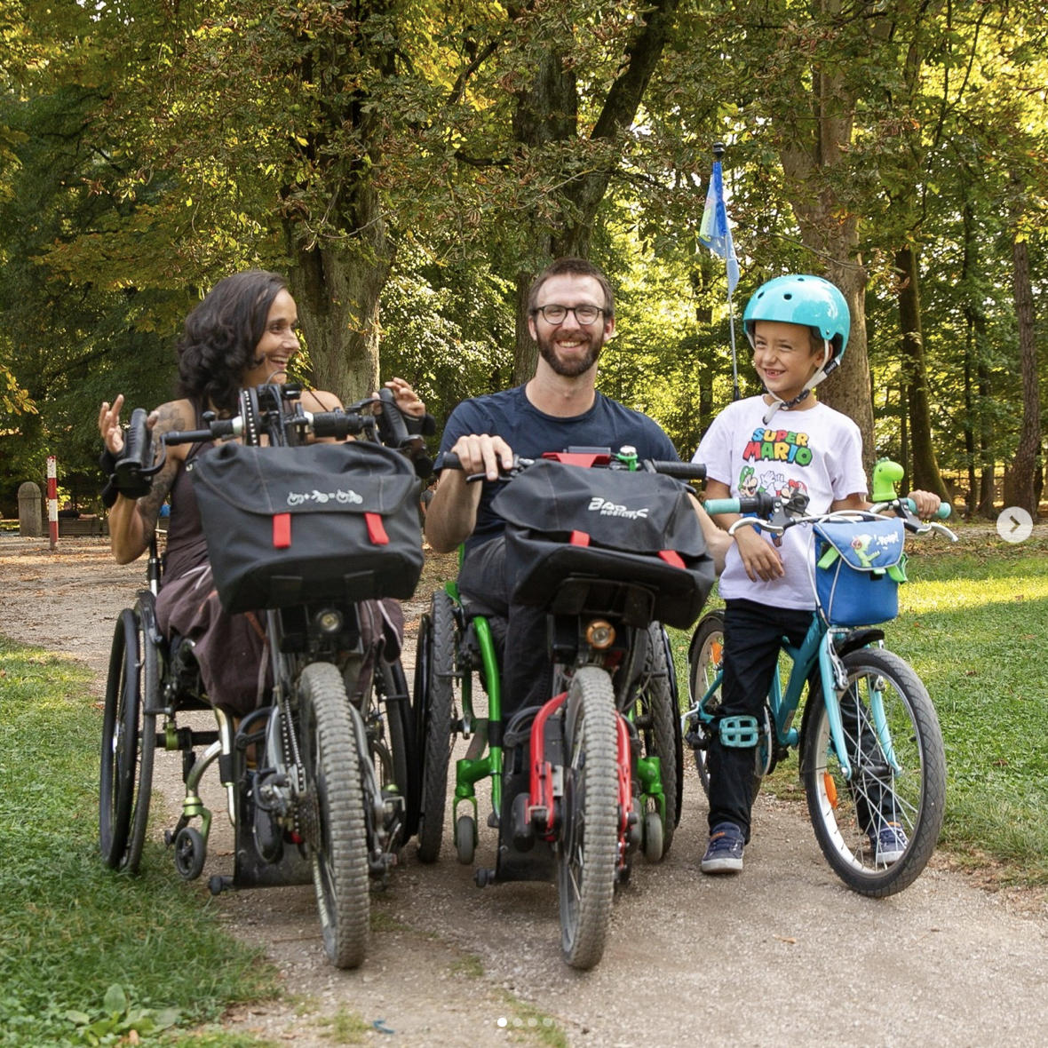 two adults and a child outdoors at a park with trees and dirt trail. Both the woman and the man are using wheelchairs that have an electric handbike attached and a large black bag on the front of each handbike. The child, is using a bike with a blue triangular flag on the back, and is wearing a blue helmet and a Super Mario t-shirt. They are all smiling. The woman has medium toned skin and shoulder length dark brown wavy hair. The man has lighter skin, short medium brown hair and beard. The child appears to be about 8 to 11 years old.
