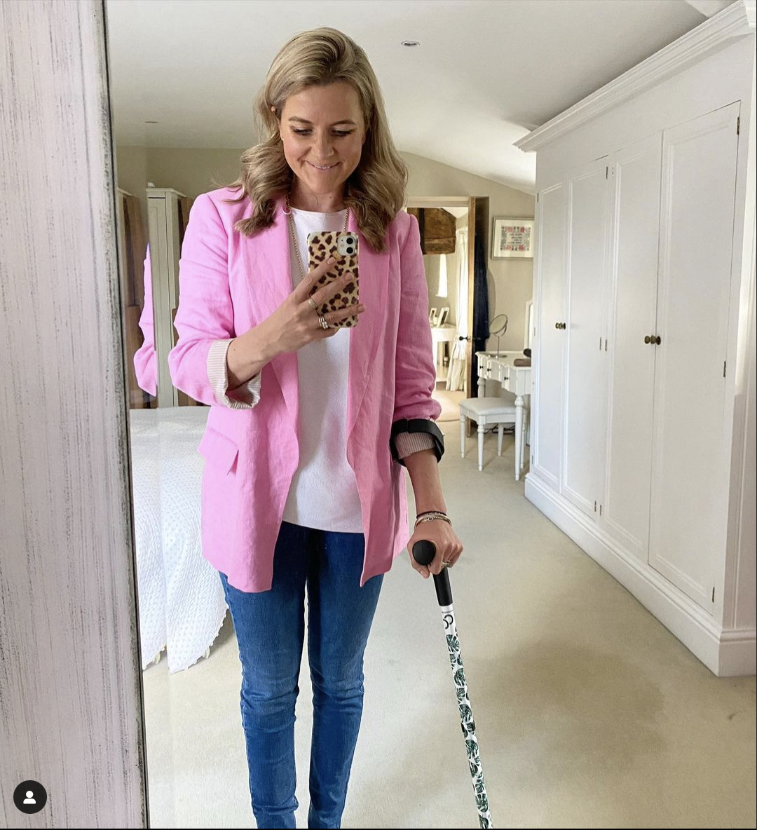 Younger middle aged white woman with shoulder length blonde hair tucked behind her ears. She's smiling while looking at her animal print covered phone held in her right hand which is the left side of the photo. She is wearing a bright pink blazer and blue jeans.  She is using a forearm crutch with her left hand and arm. The crutch has black forearm cuff and handle. The stick part is white with a green leaf design.