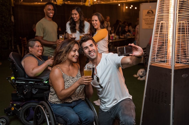 'Group of six friends at a pub all smiling and holding pint glasses up for cheets while taking a selfi photo. The two in the foreground are a man and a woman. The woman has long dark, wavy hair with golden highlights, medium skin tone, and is in an active user, low back, black manual wheelchair. She is wearing a animal print spagetti strap tank top and jeans. The man has medium brown short hair, no beard, and is wearing a tshirt and jeans. There are four people in the background. The one on the left and closest to the two in front is smiling, has dark short hair with the sides buzz cut. She's wearing a grey tank top and is using a black electric wheelchair. The other three friends are one man and two women standing up. The man is wearing a kaki green t-shirt, has short black hair buzzed shorter on the sides and has dark skin. The woman beside him is wearing a white t-shirt has long brown hair and medium toned skin. The last woman on the back right is wearing an elblow length baseball style t-shirt with a white body and yellow arms.'
