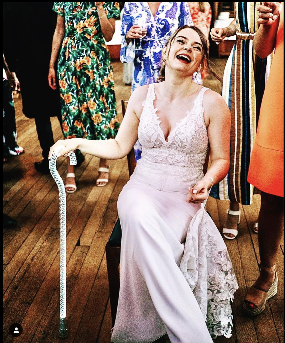 young women sitting down at a formal party or wedding reception. She's laughing, her blonde hair is in an updo and she's wearing a long lace trimmed light pink v-neck dress. She's holding a clear walking cane in her right hand. The cane has a twisted rope design and a curved handle.