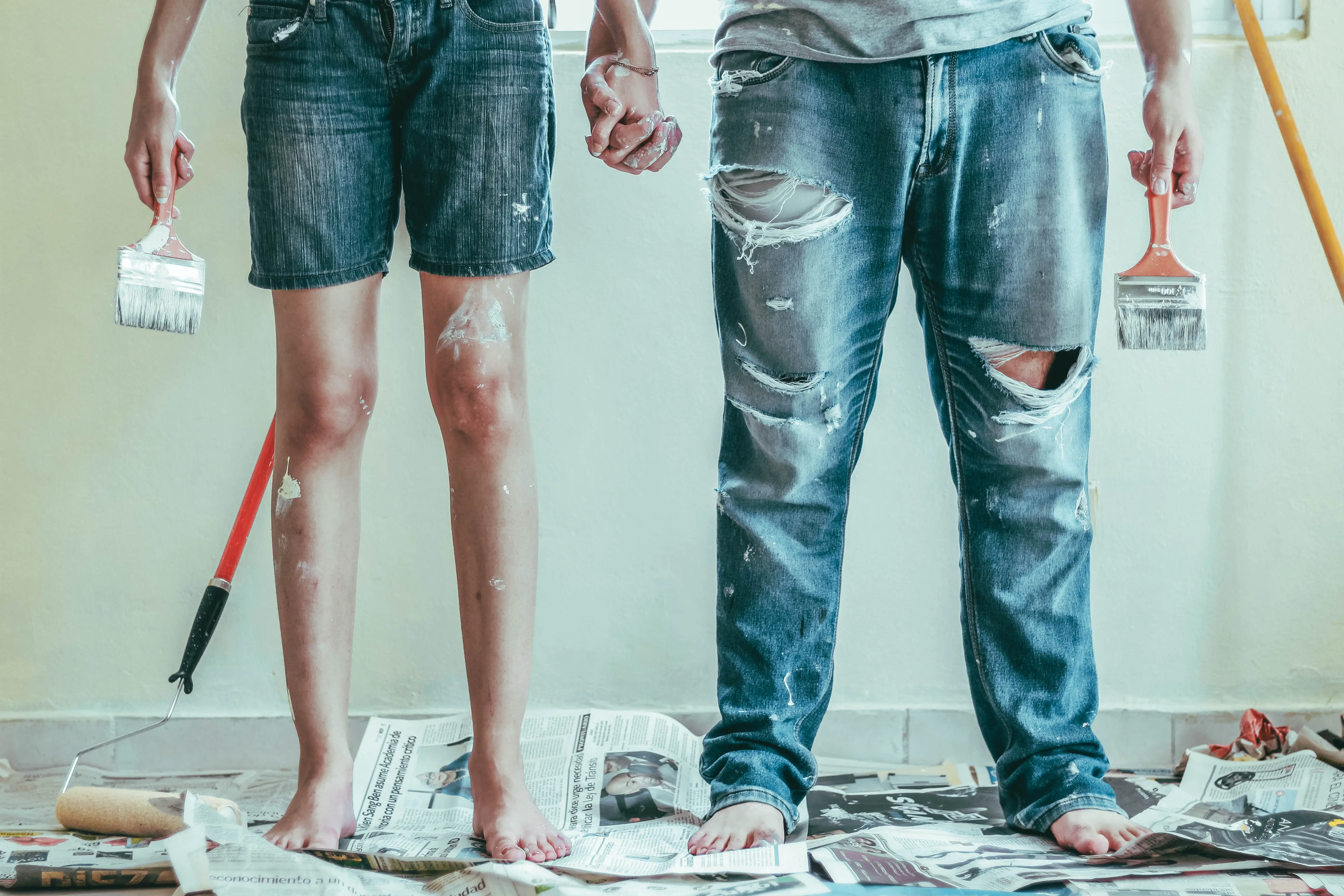 a man and a woman holding hands while holding indoor house paint brushes for painting a room. There are drips of paint all over them. The woman is wearing denim overall shorts and a white t-shirt. The man is wearing ripped blue jeans and a light grey t-shirt