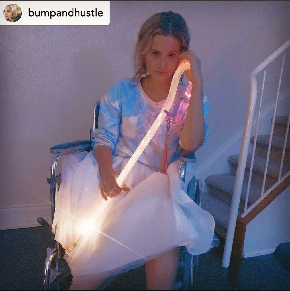 Young blonde haired woman sitting in a wheelchair at the bottom of a set of stairs in a home. She's wearing a white tulle skirt, a tie-dye top, and is holding a clear acrylic walking cane that is lit up. The upper left of the image has the instagram address @BumpandHustle.
