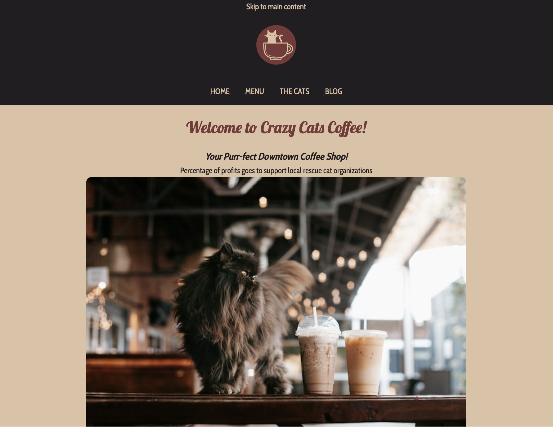 screenshot of Crazy Cats Coffee website. Colour theme is beige and brown. Top navigation is dark black-brown with beige text. It says Skip to main content. Underneath that there's the round red-brown logo of a beige cat sitting in a coffee cup. underneath there's links to HOME MENU and THE CATS. Below in red-brown calligraphy text is Welcome to Crazy Cats Coffee! The next line says in bold italic black text Your Purr-fect Downtown Coffee Shop! underneath in regular weight and styled black text is Percentage of profits goes to support local rescue cat organizations. There's a picture of a dark brown long haired cat at coffee shop on a dark wood table by two iced coffees in plastic cups. Finally below the photo, in smaller red-brown calligraphy text than the welcome message is About Crazy Cats Coffee