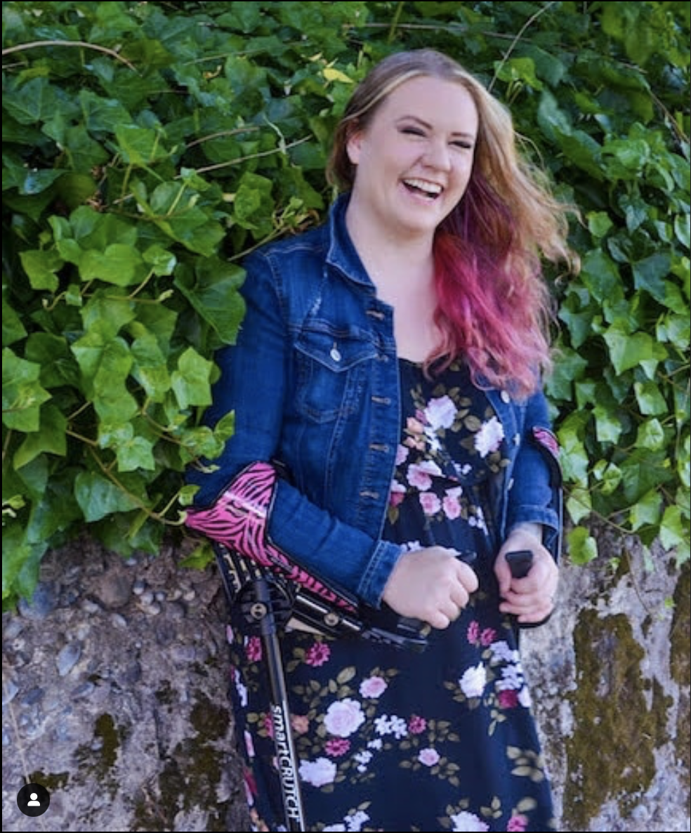Smiling young white woman with long blonde hair with bright pink dyed ends. She's wearing a medium denim jacket and  a navy dress with pink flowers on it. Her crutches have pink and black zebra stripped forearm sleeves that go around her arm for about 2cm below her elbow and all along the underneath of her lower arms. The crutches have black handles. She's outside by a stone wall covered with lichens and moss, the top of the wall is thick with ivy.